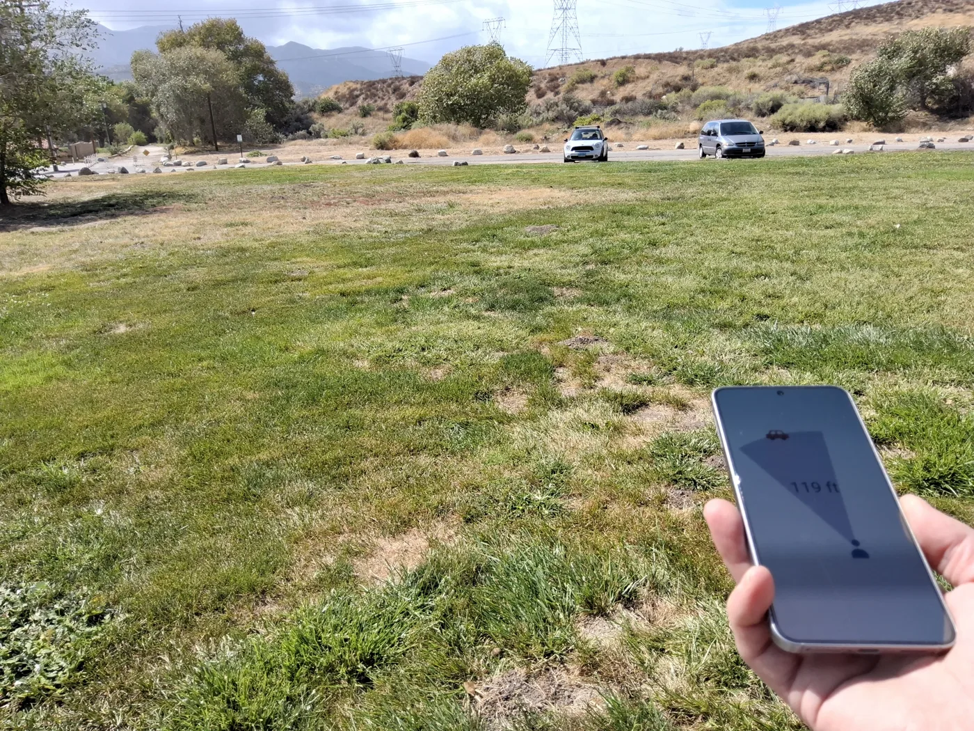 A hand holds a phone in a grassy park; the CarFinder guidance screen shows the car 119 feet away, and a parked car is visible in the distance.